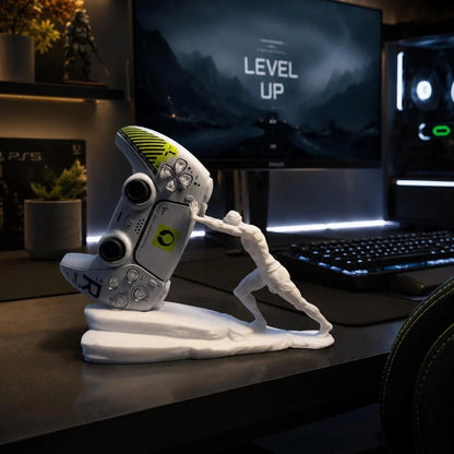 A white gaming desk accessory featuring a stylized figure pushing a PlayStation-style controller up a curved base, displayed on a desk in front of a glowing monitor and keyboard.