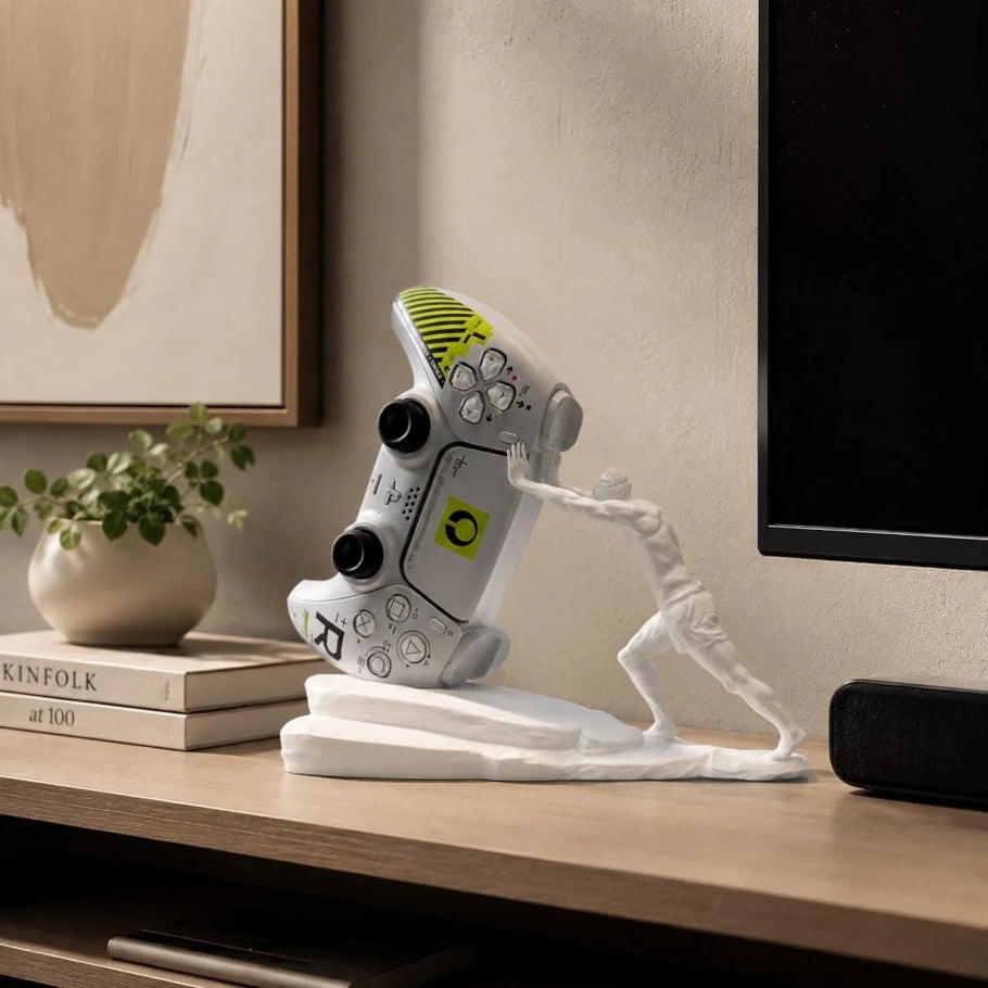 a white gaming controller sculpture displayed on a white base, pushing a small white figure. the sculpture is styled on a wooden console table next to a potted plant, books, and a television in a modern living room setting.