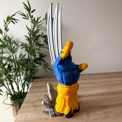 A Wolverine collectible figurine standing on a wooden tabletop, with a potted plant and neutral wall in the background. The figurine features blue and yellow armor with silver metallic claws and a gray cracked base.