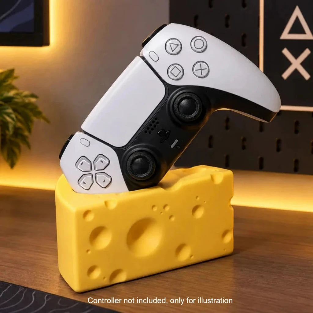 A white and black PlayStation 5 controller rests on a yellow cheese-shaped desk stand on a wooden tabletop, with warm ambient lighting and home decor in the background.