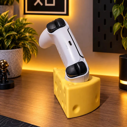 A white game controller with black buttons rests on a yellow cheese-shaped stand on a wooden desk, with warm ambient lighting and plants in the background.