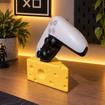 A white and black PlayStation-style controller rests on a yellow cheese-shaped desk stand on a wooden tabletop, with a framed wall sign, plants, and warm ambient lighting in the background.