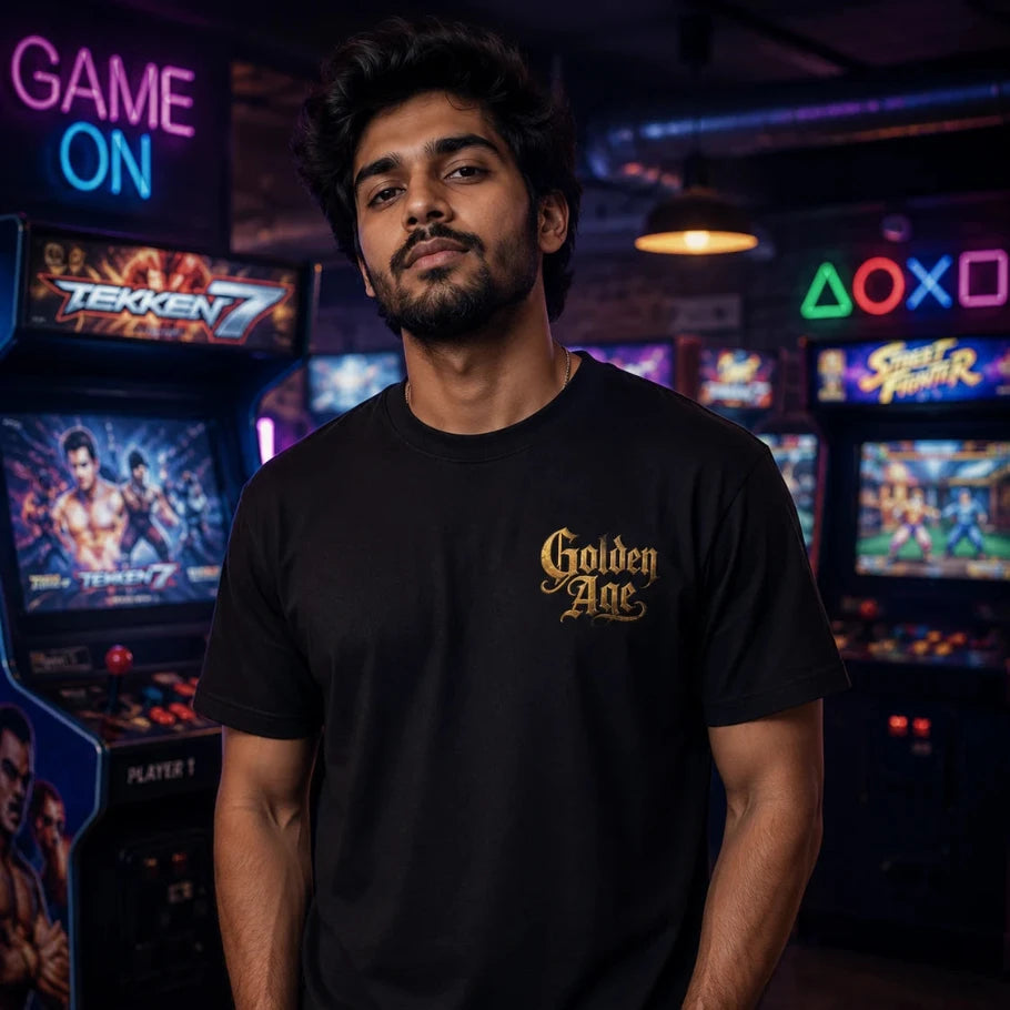 a man wearing a black t-shirt with a gold 'golden age' graphic on the left chest, standing in a dimly lit arcade room with retro game machines in the background.