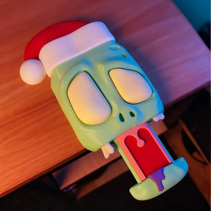 Angled view of a zombie headphone stand with a pink brain, bulging eyes, and a slime-dripping red tongue, next to a white Xbox controller on a desk.