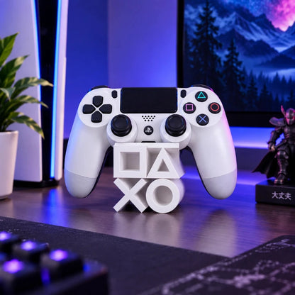 A white PlayStation 5 DualSense wireless controller stands on a white 3D letter stand spelling PS5, set on a desk in a gaming setup with blue ambient lighting, a keyboard, a plant, and a monitor in the background.