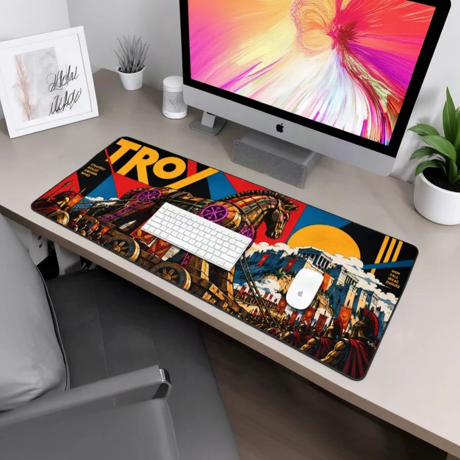 A large rectangular desk mat featuring a colorful graphic design with the word 'TROY' and an illustrated scene, shown on a desk with a keyboard, mouse, iMac, chair, and plant.