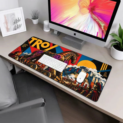 A large rectangular desk mat featuring a colorful graphic design with the word 'TROY' and an illustrated scene, shown on a desk with a keyboard, mouse, iMac, chair, and plant.