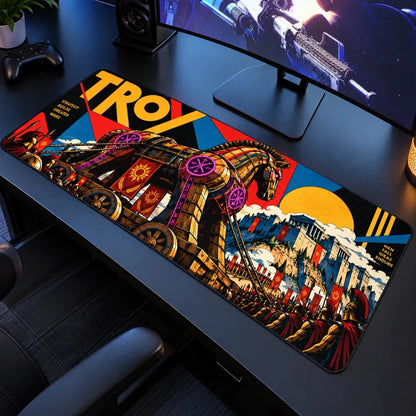 A large rectangular gaming desk mat featuring a colorful illustrated design of the Trojan Horse, with the word "TROY" in bold yellow letters, is shown on a dark desk in a computer setup. The desk mat has a complex graphic design with multiple colors including yellow, red, blue, black, and gold.