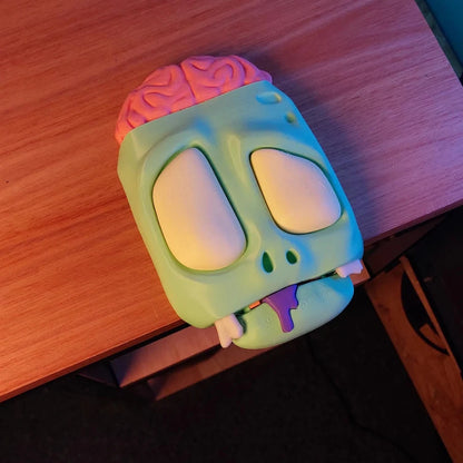 Zombie-themed headphone stand with a pink brain topper, bulging white eyes, and a mouth closed dripping purple slime, resting on a wooden desk.