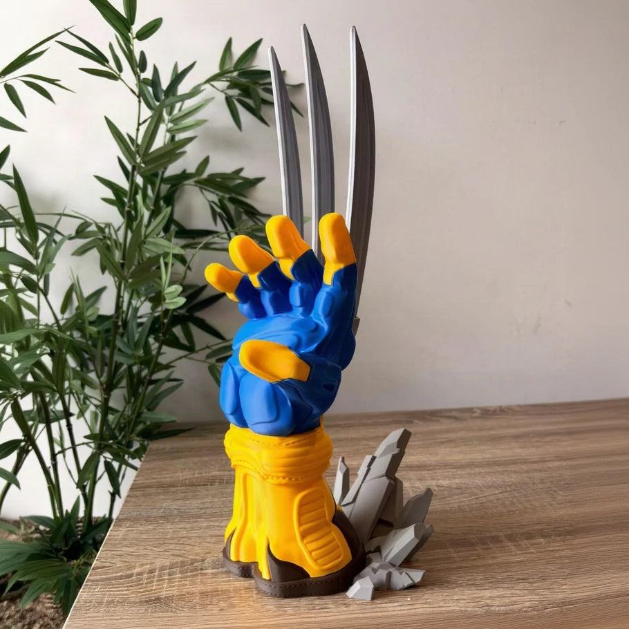 a berserker claw clawed glove hand sculpture or collectible figure standing on a wooden tabletop, with blue and yellow glove details, long silver claws, and gray cracked base pieces. a plant and beige wall are visible in the background.